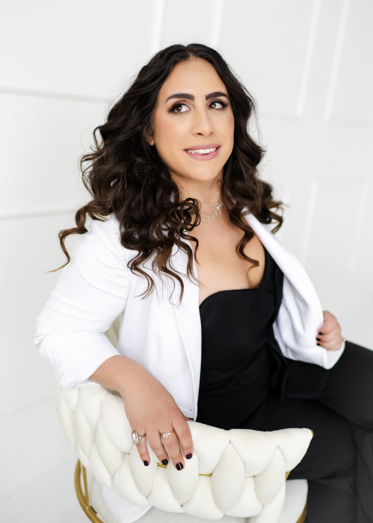 Alicia seated in a chair, wearing a white blazer, and black top and pants. She is smiling and looking up.